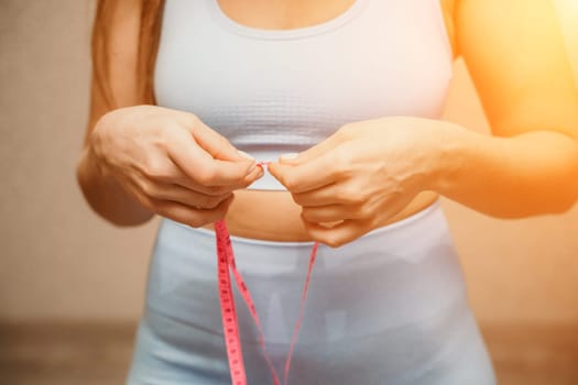 Cropped view of slim woman measuring waist with tape measure at home, close up. European woman checking the result of diet for weight loss or liposuction indoors.