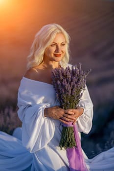 Blonde woman poses in lavender field at sunset. Happy woman in white dress holds lavender bouquet. Aromatherapy concept, lavender oil, photo session in lavender.