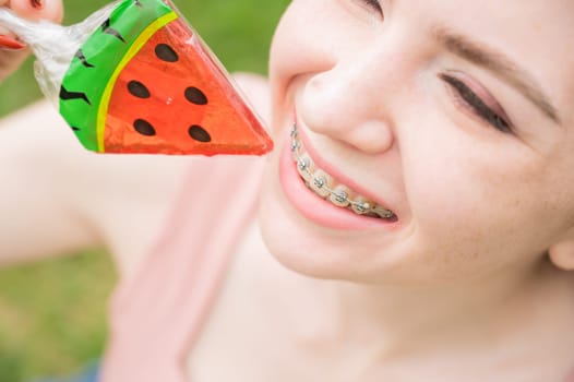 Beautiful young woman with braces on her teeth eats a watermelon-shaped lollipop outdoors