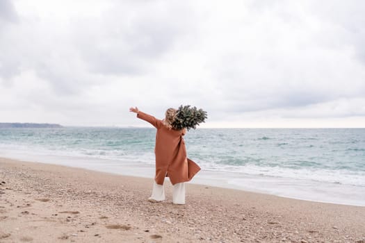 Blond woman Christmas sea. Christmas portrait of a happy woman walking along the beach and holding a Christmas tree on her shoulder. She is wearing a brown coat and a white suit