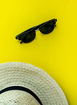 Flatlay, summer vacation. Straw hat with black ribbon and sunglasses on a yellow background. Vertical background. Background for advertising.