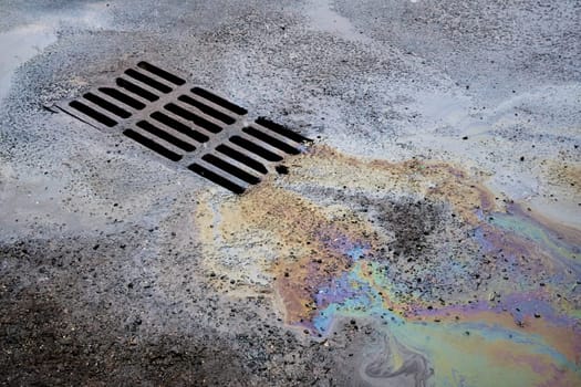 An oil or gasoline slick against the backdrop of an asphalt road flows into a storm drain. Environmental problems of water pollution.