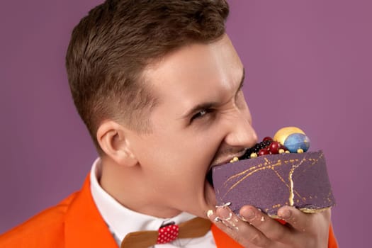 handsome birthday man in orange jacket bites yummy dessert on purple background