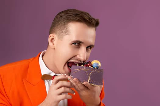 handsome birthday man in orange jacket bites cake on purple background