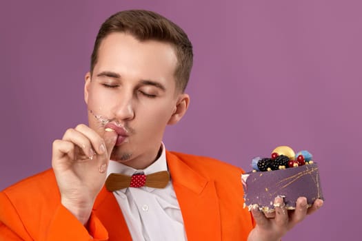 handsome man in orange jacket eating tasty cake on purple background. celebrating birthday, enjoying holidays tasty food