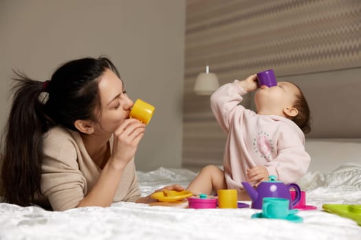 happy mother and little child daughter pretending drinking tea from plastic toy cups in bedroom, family having fun