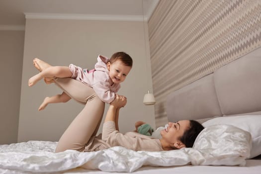 Happy Caucasian mother raising laughing cute baby and playing together in bedroom