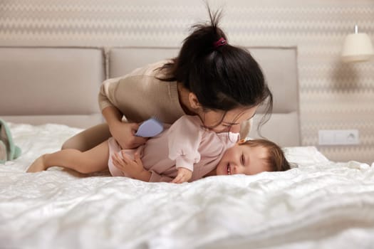 happy mother playing with her cute baby daughter on bed in bedroom. loving family having fun