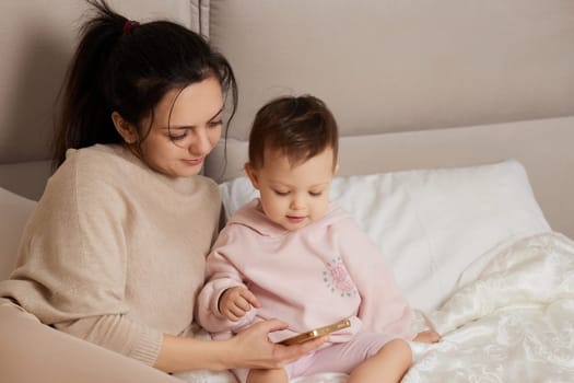 Mother and child playing with smartphone while sitting on bed in bedroom at home