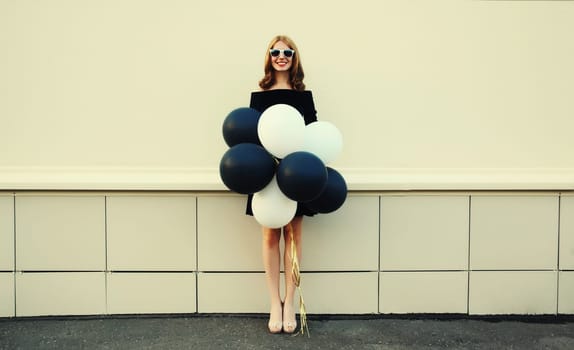 Beautiful happy smiling young woman full length with bunch of black and white balloons in summer dress on city street