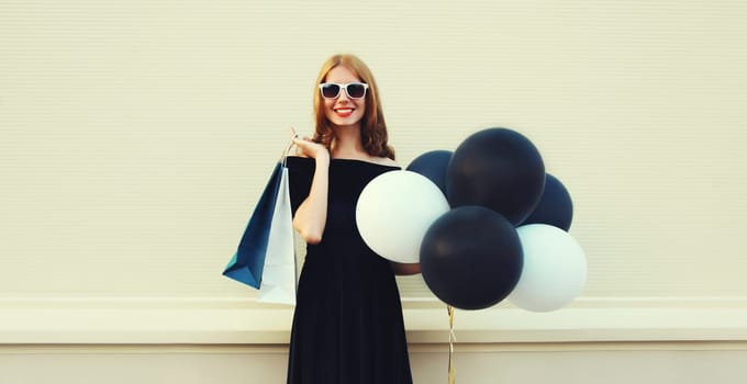 Beautiful happy smiling young woman with shopping bags and bunch of black and white balloons in summer dress on city street