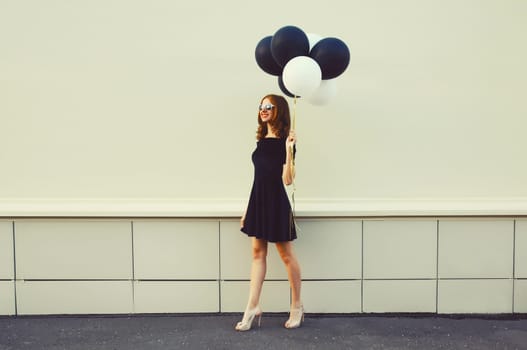 Beautiful happy smiling young woman full length with bunch of black and white balloons in summer dress on city street