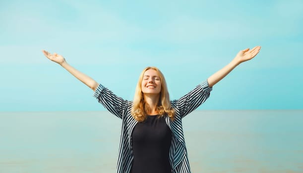 Summer vacation, happy young woman raising her hands up on the beach on sea coast and blue sky background