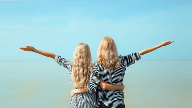 Summer vacation of two women raises her hands up on the beach, happy caucasian girlfriends or mother with adult daughter together on sea coast and blue sky background