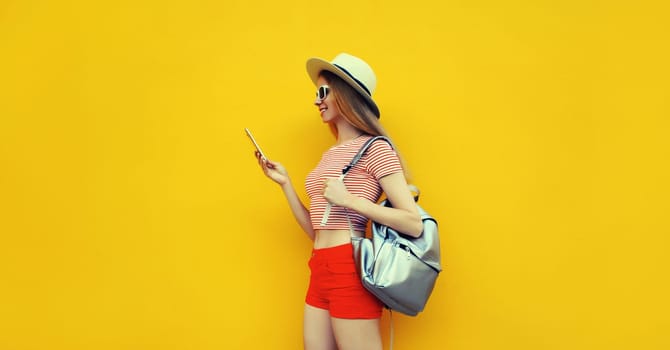Summer portrait of happy traveler young woman 20s with mobile phone looking at device in casual straw hat with backpack on bright yellow studio background