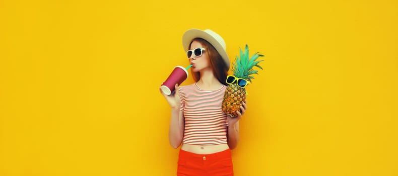 Summer bright portrait of stylish young woman drinking fresh juice holding pineapple fruit in straw hat, sunglasses on colorful yellow studio background