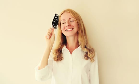 Portrait of happy smiling caucasian blonde young woman combing her hair with comb on white studio background