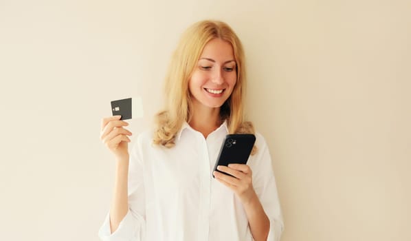 Portrait of happy smiling young woman holding plastic credit bank card, looking at smartphone, shopping online or ordering goods