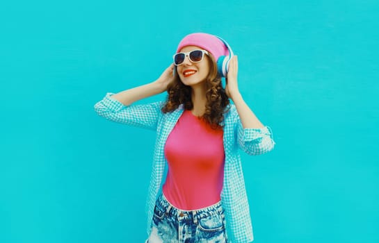 Portrait of stylish modern happy young woman listening to music with headphones wearing colorful pink hat on blue studio background