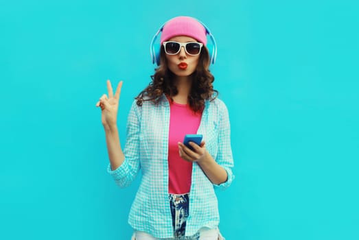 Portrait of stylish modern happy young woman listening to music in headphones with mobile phone wearing pink hat, sunglasses on blue studio background