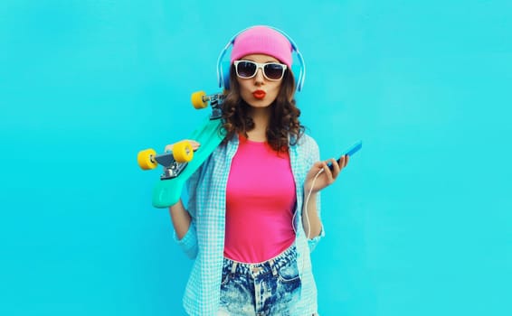 Portrait of stylish modern happy young woman listening to music in headphones with mobile phone and skateboard posing wearing colorful clothes, pink hat, sunglasses on blue studio background