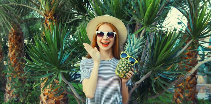 Summer portrait of happy smiling young woman with pineapple fruits wearing sunglasses, straw hat posing on palm tree background