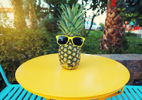 Summer image of stylish pineapple fruit in yellow sunglasses on the table in street cafe in city park