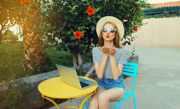 Happy cool young woman blowing her lips sends kiss with laptop sitting at the table in street cafe in summer park