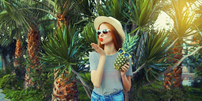Summer portrait of beautiful young woman with pineapple fruits posing blowing her lips sends kiss wearing sunglasses, straw hat on palm tree background