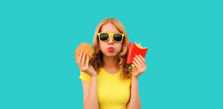 Portrait of happy cheerful young woman eating burger fast food and french fries, fried potatoes isolated on blue studio background