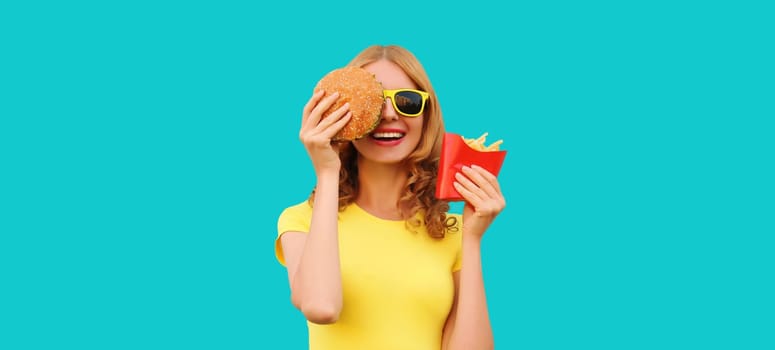 Portrait of happy cheerful young woman eating burger fast food and french fries, fried potatoes isolated on blue studio background