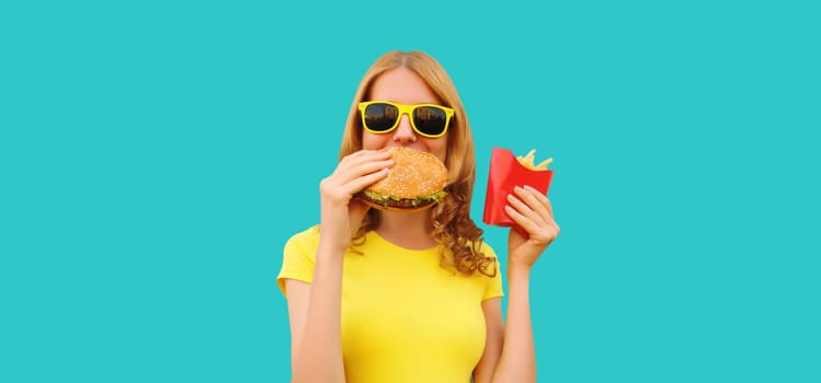 Portrait of happy cheerful young woman eating burger fast food and french fries, fried potatoes isolated on blue studio background