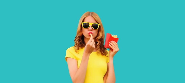 Portrait of stylish young woman eating french fries, fried potatoes fast food in sunglasses on blue studio background