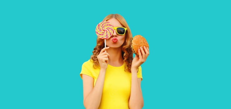 Portrait of happy cheerful young woman having fun with burger fast food and colorful sweet lollipop isolated on blue studio background