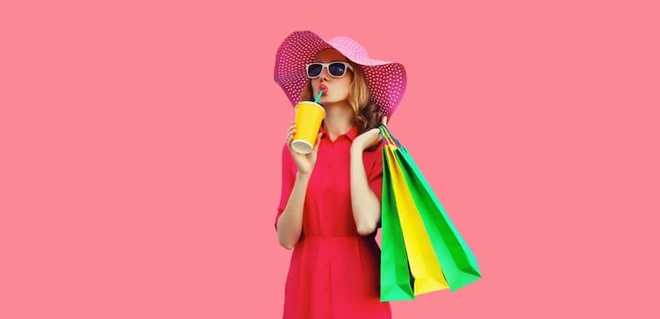Portrait of beautiful relaxed young woman model with colorful shopping bags drinking juice in summer straw hat, dress on pink studio background