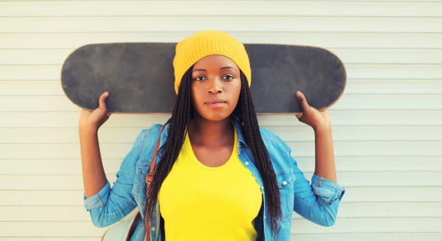 Portrait of modern young african woman model posing in colorful clothes, yellow hat with skateboard in the city