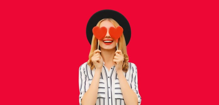 Portrait of happy cheerful young woman with sweet red heart shaped lollipop on stick in black round hat on pink studio background