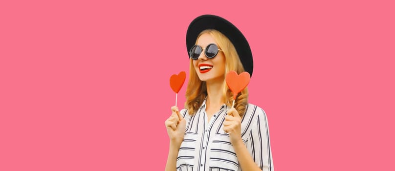 Portrait of happy cheerful young woman with sweet red heart shaped lollipop on stick in black round hat on pink studio background