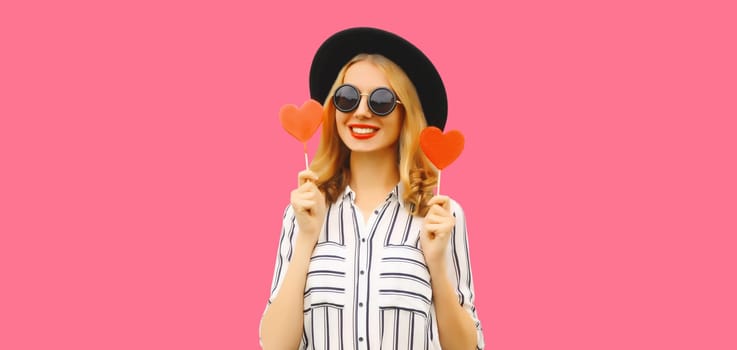 Portrait of happy cheerful young woman with sweet red heart shaped lollipop on stick in black round hat on pink studio background