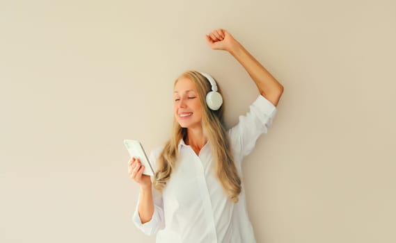 Happy cheerful middle aged woman listening to music with mobile phone in headphones having fun dancing on studio background