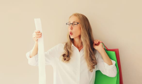 Shocked and surprised caucasian middle aged woman in eyeglasses looks amazed at the high price tag on long paper receipt holding shopping bag