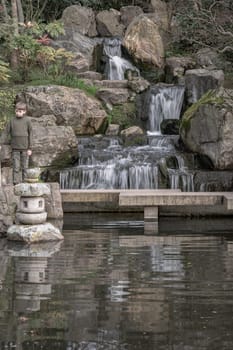 London, UK - Feb 15, 2024 - A boy stands on a stone bridge in front of waterfall and rocky cliffs in Kyoto garden, Space for text, Selective focus.