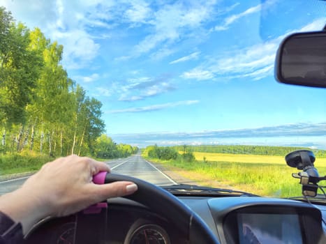 Car salon, windshield, hand of woman on steering wheel and landscape. View from seat of driver on nature with Road, trees, blue sky at a sunny day. Single trip of female traveler. Partial focus