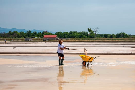 Nin, Croatia - 8 July 2021: Man picking up salt in the swamp and filling the wheelbarrow. Salt farmer harvesting the salt. Sea salt is salt that is produced by the evaporation of seawater.