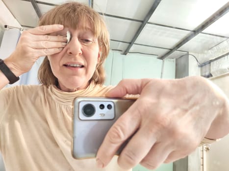 Woman removing eye makeup with a napkin. A cheerful middle aged lady capturing playful selfie using her smartphone in front of a bathroom mirror