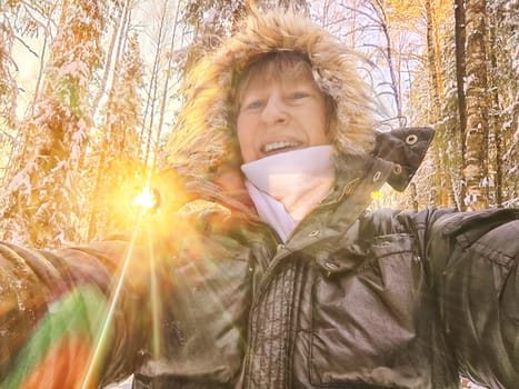 A cheerful middle aged woman in a winter coat with fur, scarf taking selfie outdoors on nature in a forest or park in beautiful sunny day