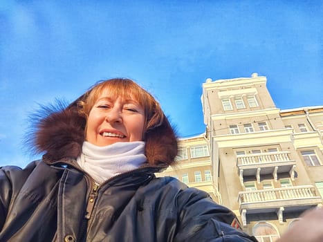 A cheerful woman poses taking selfie in front of an elegant, multi-storied building under a clear blue sky in winter, autumn or spring