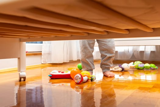 Baby standing near a crib with toys on the floor. View from under the bed. The window brings in
