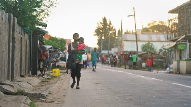 Ranohira, Madagascar - April 29, 2019:  Unknown Malagasy woman carrying baby kid, walking on asphalt road at small African city, more blurred people in background, late afternoon scene