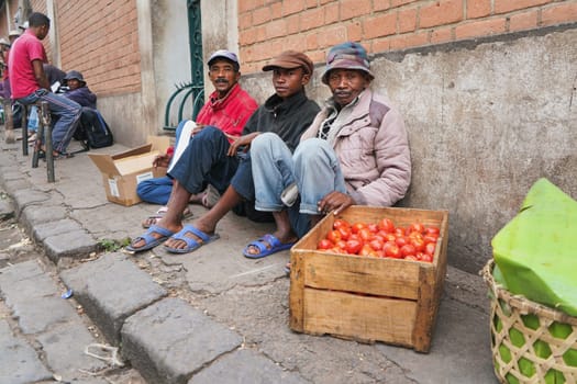 Antananarivo, Madagascar - April 24, 2019: Three unknown Malagasy men sitting on sidewalk, selling tomatoes. Food is usually sold on streets in Madagascar